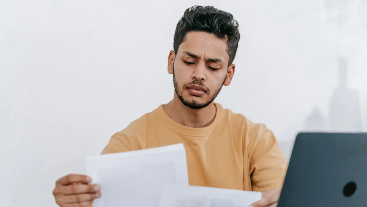 Mann hält zwei Blätter Papier nebeneinander und vergleicht sie, daneben ein Laptop