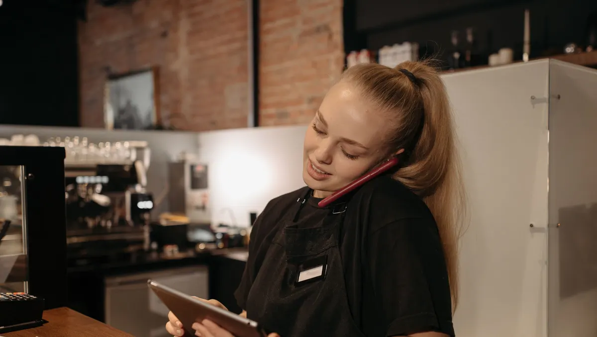 Frau in Schürze steht in einem Café am Tresen, telefoniert und schaut auf ein Tablet
