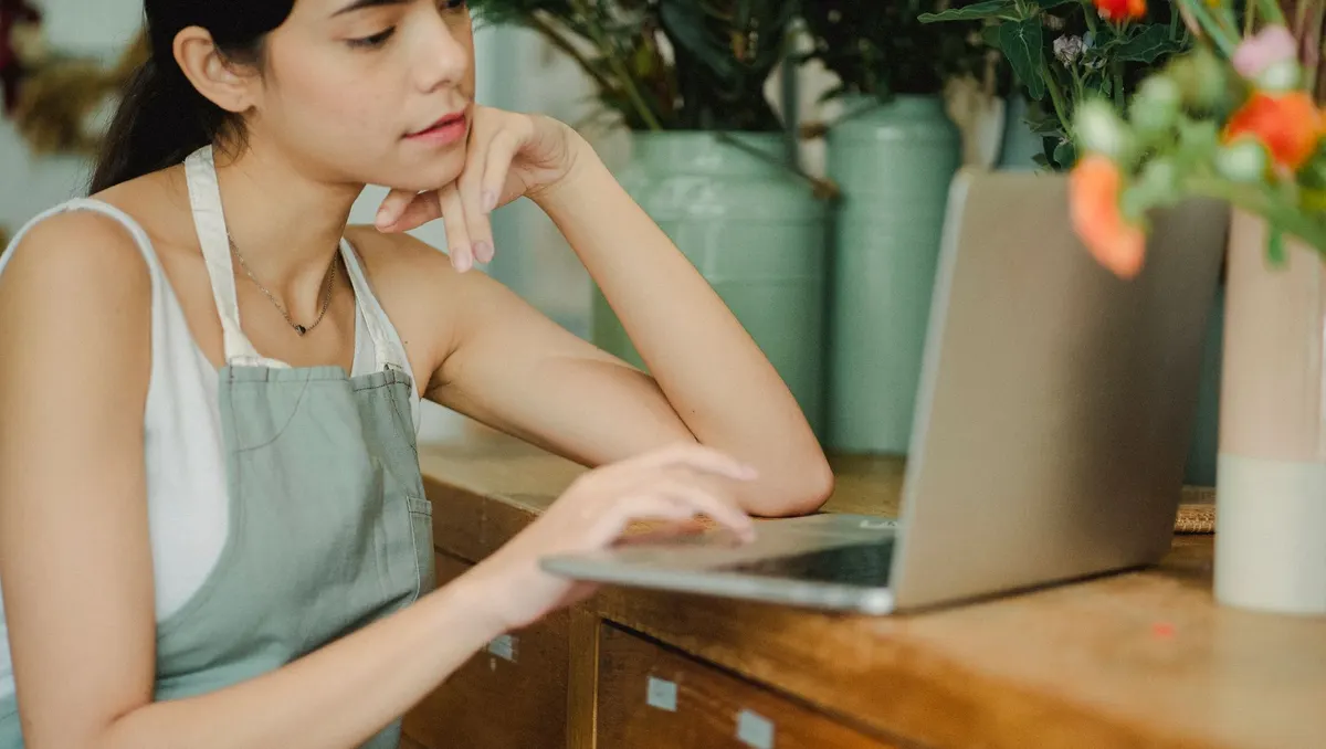Frau in Schürze sitzt in einem Blumenladen am Holztresen vor einem Laptop und überlegt, das Kinn auf die Hand gestützt